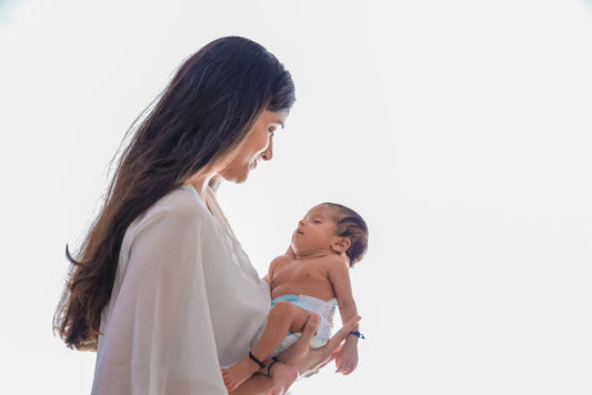 New mother holding her infant while wearing a delicate gold pendant necklace, soft natural lighting, intimate editorial portrait