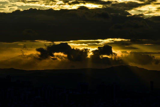 Golden sunbeams burst through dramatic clouds above a silhouetted city skyline at sunset